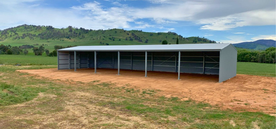 Open-front steel farm shed on a rural property, set against green hills and blue sky